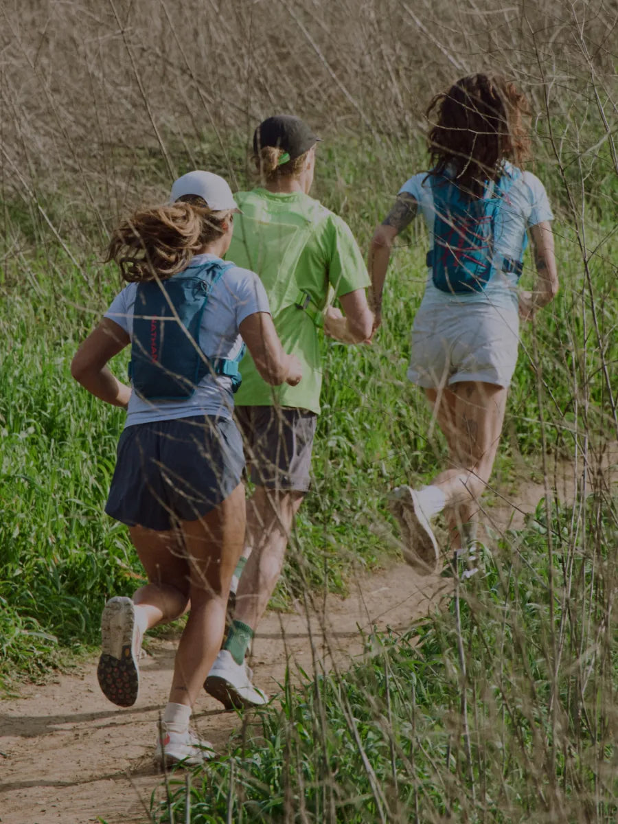 Three trail runners are seen from behind running on a dirt path through tall grass, wearing NATHAN hydration packs and athletic clothing.