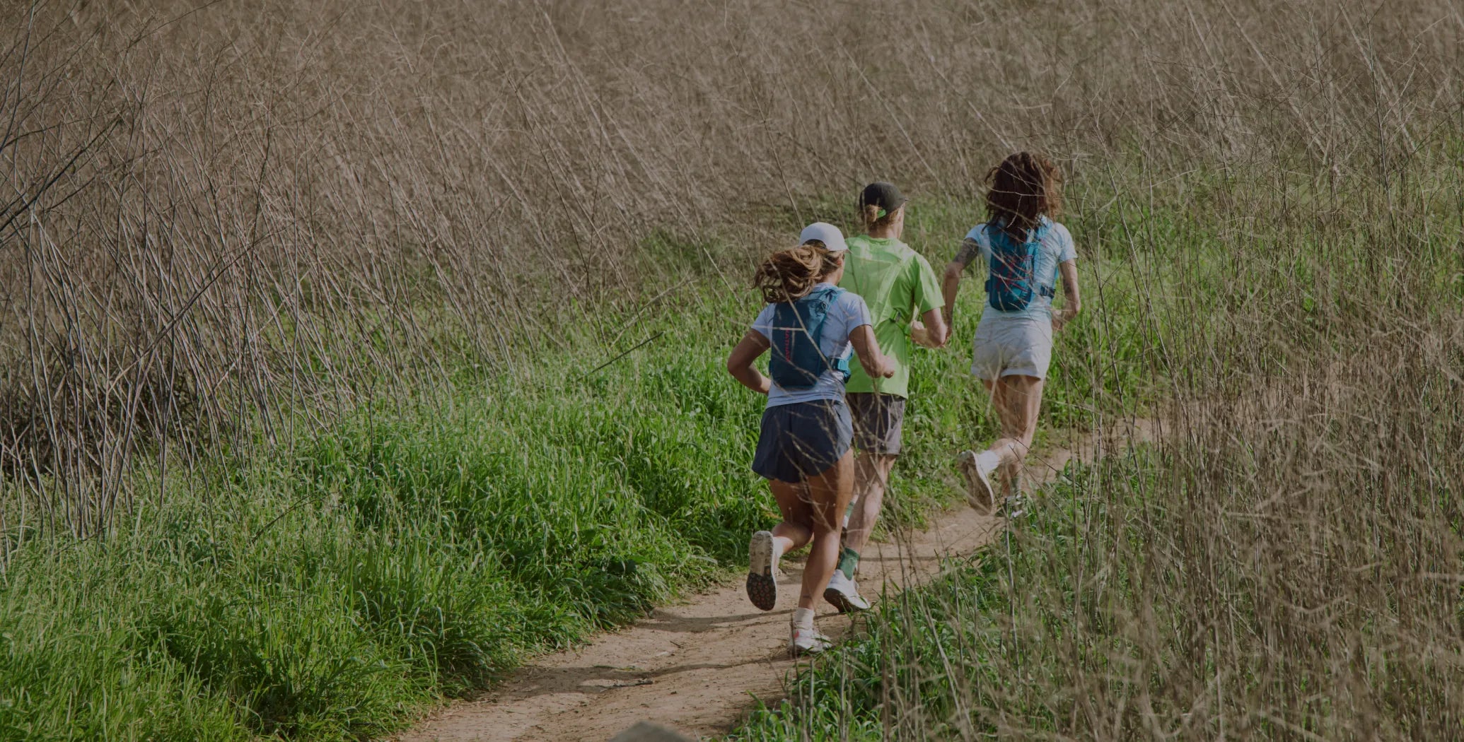 Three trail runners are seen from behind running on a dirt path through tall grass, wearing NATHAN hydration packs and athletic clothing.