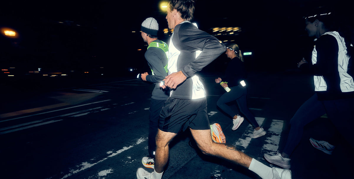 Three trail runners are seen from behind running on a dirt path through tall grass, wearing NATHAN hydration packs and athletic clothing.