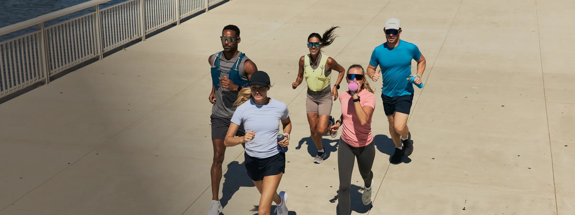 Five runners jogging together on a sunny outdoor boardwalk or pier, wearing NATHAN hydration vests and carrying NATHAN handheld flasks.