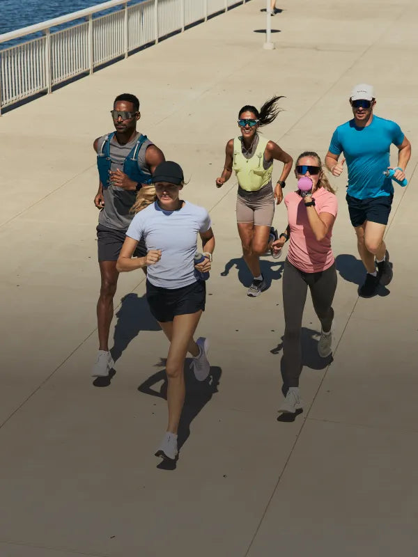Five runners jogging together on a sunny outdoor boardwalk or pier, wearing NATHAN hydration vests and carrying NATHAN handheld flasks.